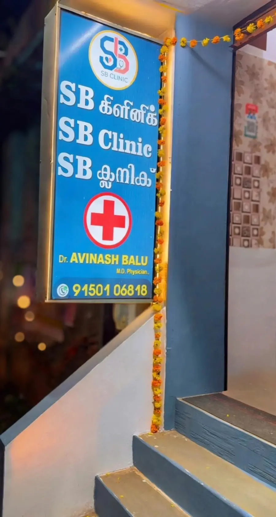 The lit SB Clinic signboard beside the entrance steps, with the doctor's name and phone number